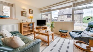 A cozy living room with a green sofa, a wooden coffee table, a TV, and a large window with a view of a stone building. - 1 Bed in Reynoldston (oc-hh508) (Scurlage)