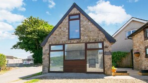 A stone cottage with a large window and a wooden door, surrounded by gravel and a tree. - 1 Bed in Reynoldston (oc-hh508) (Scurlage)