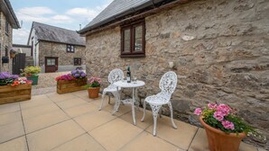 A stone building with a tiled patio, white wicker chairs, and flower planters. - 1 Bed in Reynoldston (oc-hh508) (Scurlage)