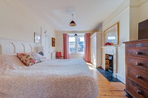 A bedroom with a large bed, a dresser, a chair, a fireplace, and a window with curtains. - 5 Bed in Southwold (oc-sfamo) (Southwold)
