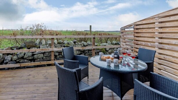 A round glass table with wicker chairs on a wooden deck, surrounded by a stone wall and wooden fence, overlooking a grassy field. - 2 Bed in Alnwick (oc-80499) (Newton-by-the-Sea)