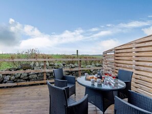 A round glass table with wicker chairs on a wooden deck, surrounded by a stone wall and wooden fence, overlooking a grassy field. - 2 Bed in Alnwick (oc-80499) (Newton-by-the-Sea)