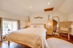 A bedroom with a large bed, wooden flooring, a rocking chair, and a view of the outdoors.