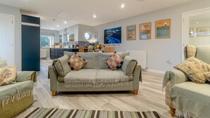 A modern living room with a sofa, two armchairs, a dining area, kitchen, and a large window. - 2 Bed in Snape (oc-sfvin) (Church Common)