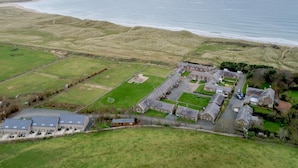 Vue aérienne d’une propriété située sur la côte, comprenant plusieurs bâtiments, un grand champ herbeux et une plage en arrière-plan.