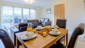 Un coin repas avec une table en bois mise pour le petit-déjeuner, comprenant des croissants et du jus d’orange. On peut voir deux chaises en cuir noir et un canapé à l’arrière-plan.
