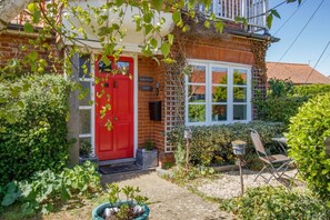 Ein Ziegelhaus mit roter Tür, einem weißen Fenster und einem Garten mit Pflanzen und einem Stuhl.
