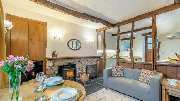 A cozy living room with a stone fireplace, wooden beams, a dining table set with plates and glasses, and a grey sofa with patterned cushions. - 2 Bed in Lindale (oc-c34128) (Lindale)