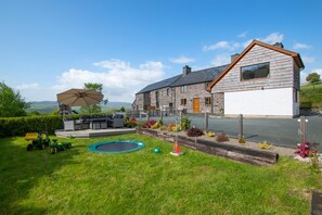 Une maison avec une façade en pierre, un garage et une pelouse aménagée avec du mobilier d’extérieur et un trampoline.