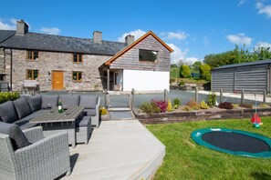 Une maison en pierre avec une extension en bois, une terrasse avec des meubles d’extérieur et un trampoline.