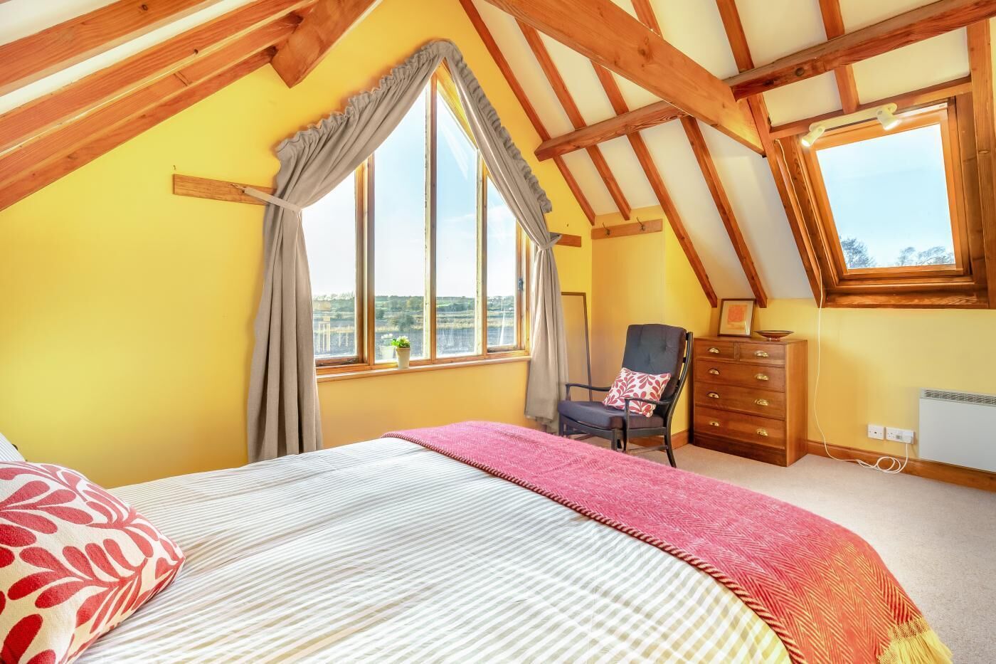 A bedroom with a sloped ceiling, a bed with a patterned pillow, a chair, a dresser, and a window with a view.