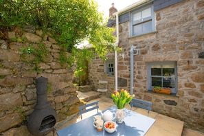 A stone building with a chimney, a table with a vase of flowers, and a window with a plant. - 3 Bed in St. Just (oc-p00674) (Carnyorth)