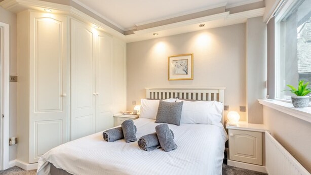 A neatly made bed with pillows and rolled towels, a bedside table with a lamp, a framed picture on the wall, and a window with a plant. - 2 Bed in Grasmere (oc-ld175) (Grasmere)