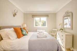 Un dormitorio con cama, almohadas, cómoda, una silla, ventana y espejo montado en la pared.