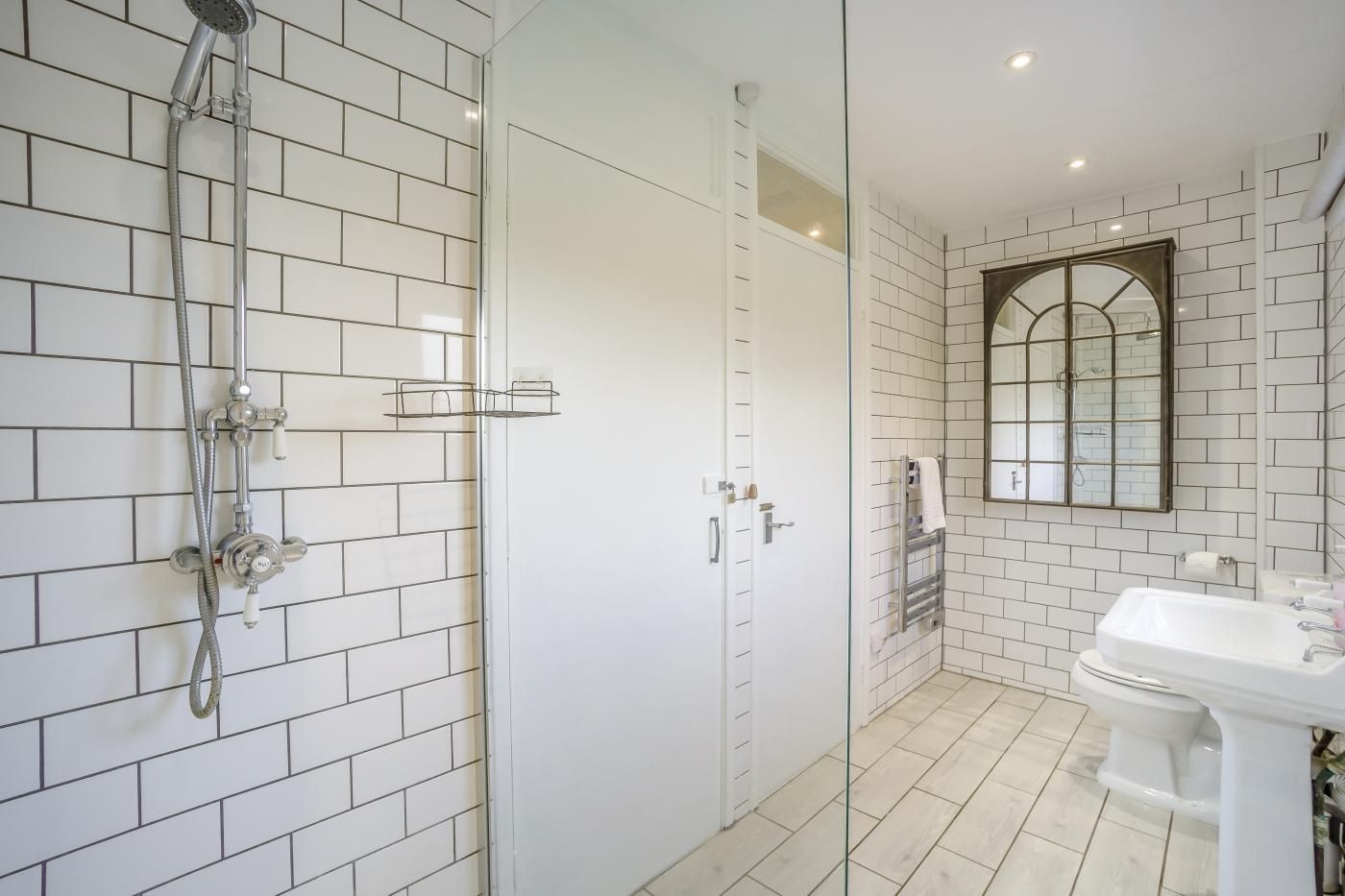A bathroom with white tiled walls, a shower with a handheld showerhead, a toilet, and a sink.