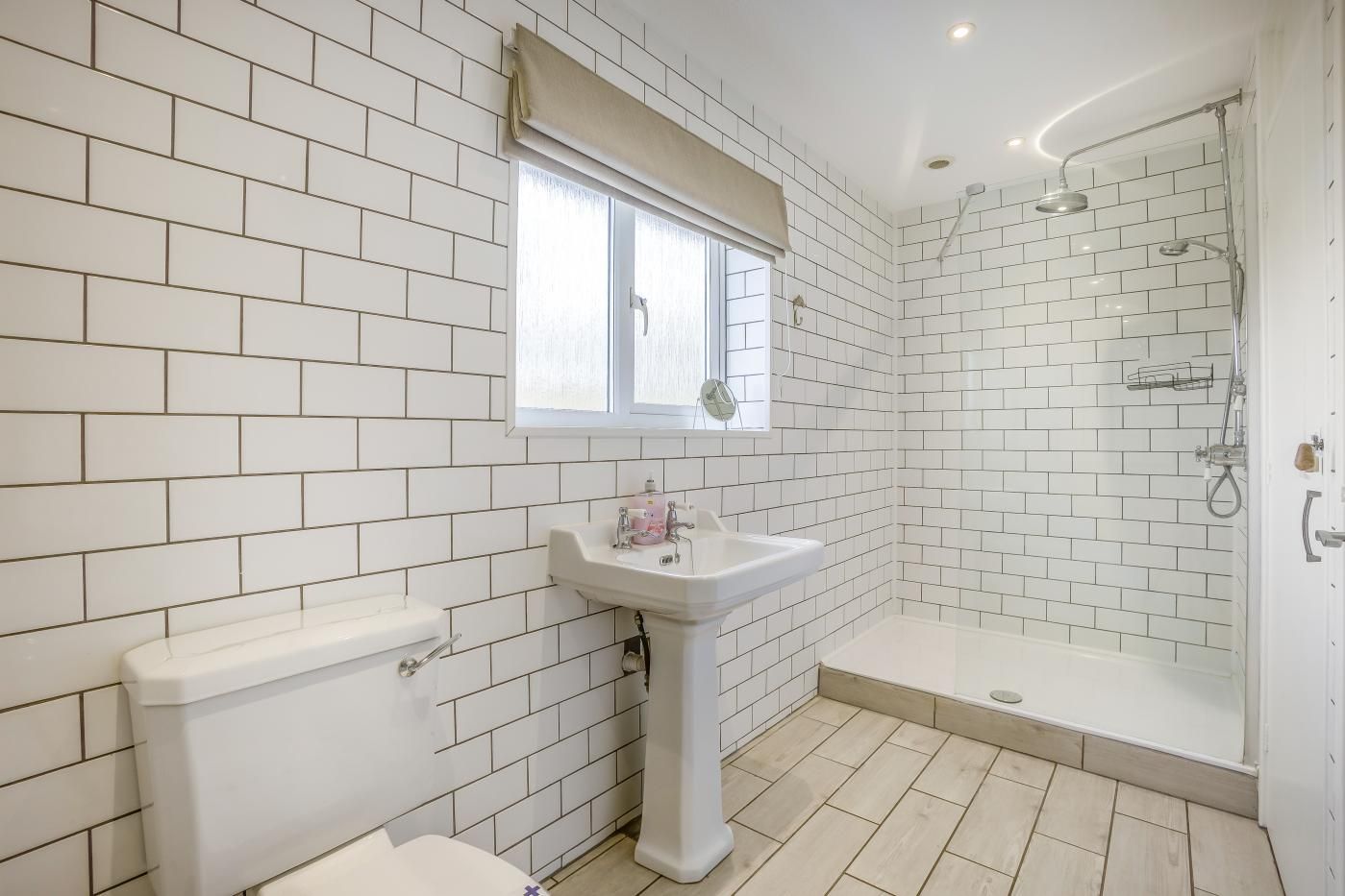 A bathroom with white tiled walls, a window, a sink, a toilet, and a shower.