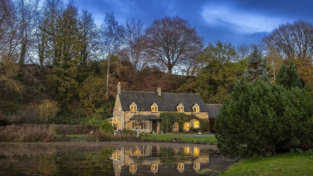 Een stenen huis met een vijver ervoor, omgeven door bomen en een heldere lucht.