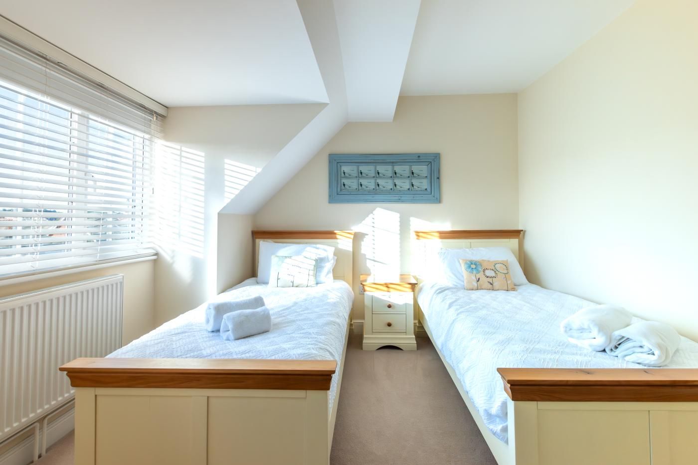 A bedroom with two single beds, a radiator, a window with blinds, and a wall-mounted light switch panel.