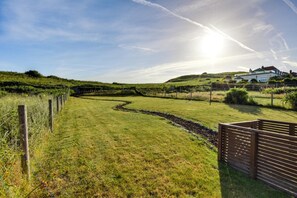 Ein grasbewachsener Pfad führt zu einem Haus, an einer Seite eine Ziegelmauer und auf der anderen ein Holztor.
