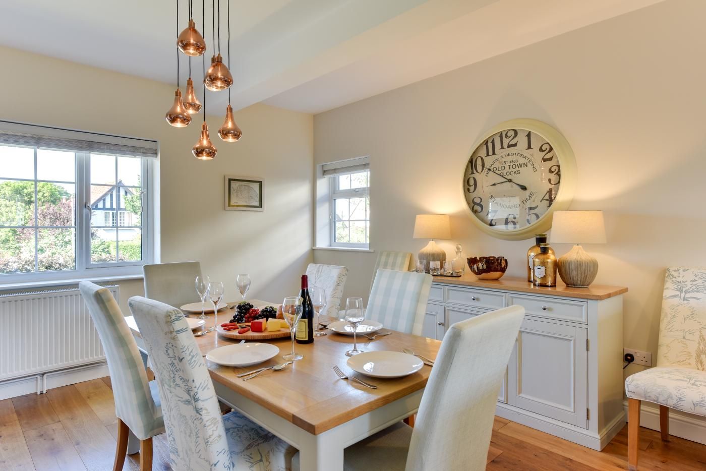A dining room with a wooden table set for four, surrounded by chairs. A large wall clock and a window with blinds are visible.