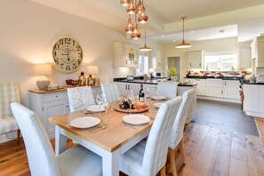 A modern kitchen with white cabinetry, a dining area with a wooden table set for six, and a large wall clock. - 4 Bed in Sheringham (oc-2330) (Sheringham)