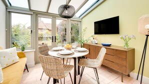 A dining area with a round table, wicker chairs, a wooden cabinet, and a television on the wall. - 2 Bed in Castle Acre (oc-2307) (Castle Acre)