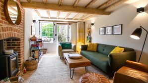 A cozy living room with a fireplace, a green sofa, a wooden ceiling, and a view through a large window. - 2 Bed in Castle Acre (oc-2307) (Castle Acre)