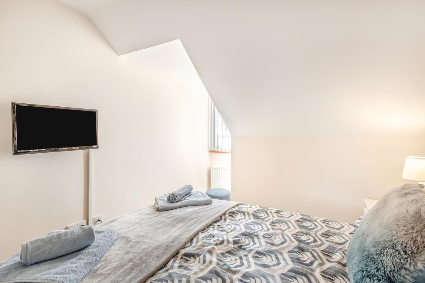 A bedroom with a bed, a flat-screen TV mounted on the wall, a window, and a lamp.