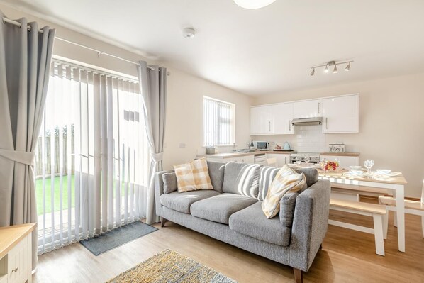 A modern living room with a grey sofa, a dining table set for two, and a kitchen in the background. - 3 Bed in Chapel St. Leonards (oc-r34145) (Chapel Saint Leonards)