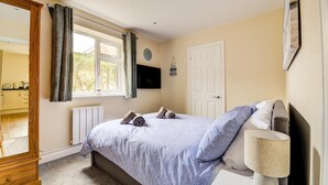 A bedroom with a bed, a bedside table, a lamp, a TV, a window with curtains, and a door. - 1 Bed in Sheringham (oc-1845) (Sheringham)