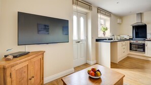 A modern kitchen with a flat-screen TV, wooden cabinets, a dining table with fruit, and a large window. - 1 Bed in Sheringham (oc-1845) (Sheringham)