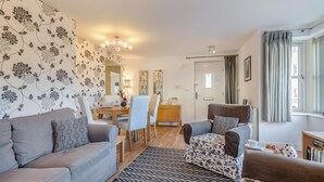 Uma sala de estar moderna com um sofá cinzento, uma poltrona estampada e um espaço de jantar com mesa e cadeiras de madeira.