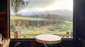 Habitació amb una finestra gran que dona a un paisatge amb gespa, una taula rodona de fusta i dues cadires.