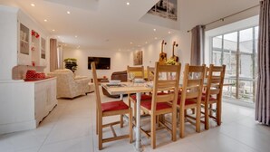 Un comedor con una mesa y sillas de madera, una zona de cocina con armarios blancos y un ventanal con cortinas.