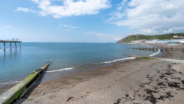 Una playa costera con un muelle que se adentra en el mar, una orilla arenosa y edificios al fondo.