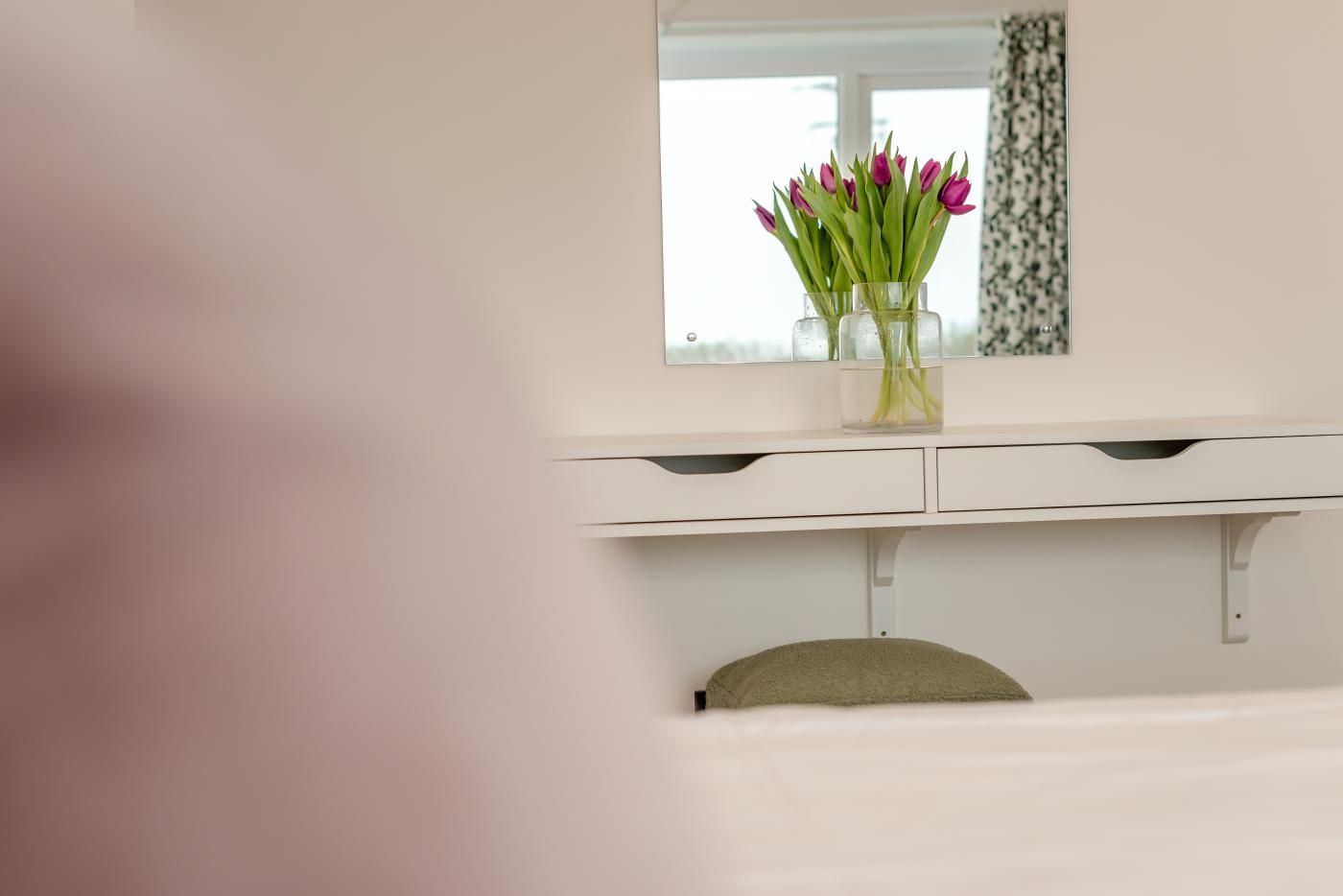 Un mobile lavabo bianco con uno specchio sopra, un vaso con fiori viola e una sedia beige sottostante.