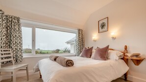 A bedroom with a large bed, a chair, a bedside table with a book, and a window with curtains. - 1 Bed in St. Merryn (oc-p00645) (Saint Merryn)