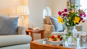 A cozy living room with a sofa, a coffee table with a floral arrangement, a lamp, and a window with curtains. - 1 Bed in St. Merryn (oc-p00645) (Saint Merryn)