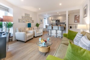 Ein modernes Wohnzimmer mit einem Glastisch, zwei weißen Sofas, einem grünen Sessel und einer Küche im Hintergrund.