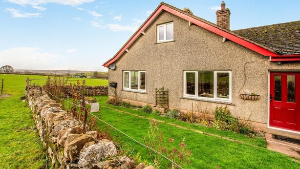 A stone house with a red door and windows, surrounded by a stone wall and a grassy field. - 2 Bed in Renwick (oc-h34195) (Renwick)