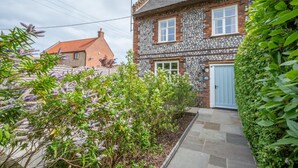 A stone house with a blue door, surrounded by lush greenery and flowering plants. - 4 Bed in Northrepps (oc-2332) (Northrepps)