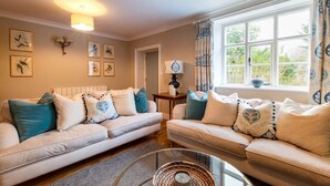 A living room with two sofas, a coffee table, and a lamp. There are framed pictures on the wall and a window with curtains. - 4 Bed in Northrepps (oc-2332) (Northrepps)