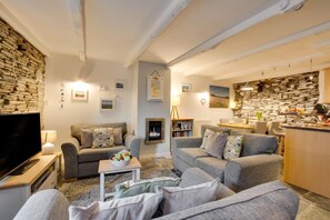 A cozy living room with a fireplace, stone wall, and a kitchen area with a stone backsplash. - 2 Bed in Padstow (oc-p00670) (Padstow)
