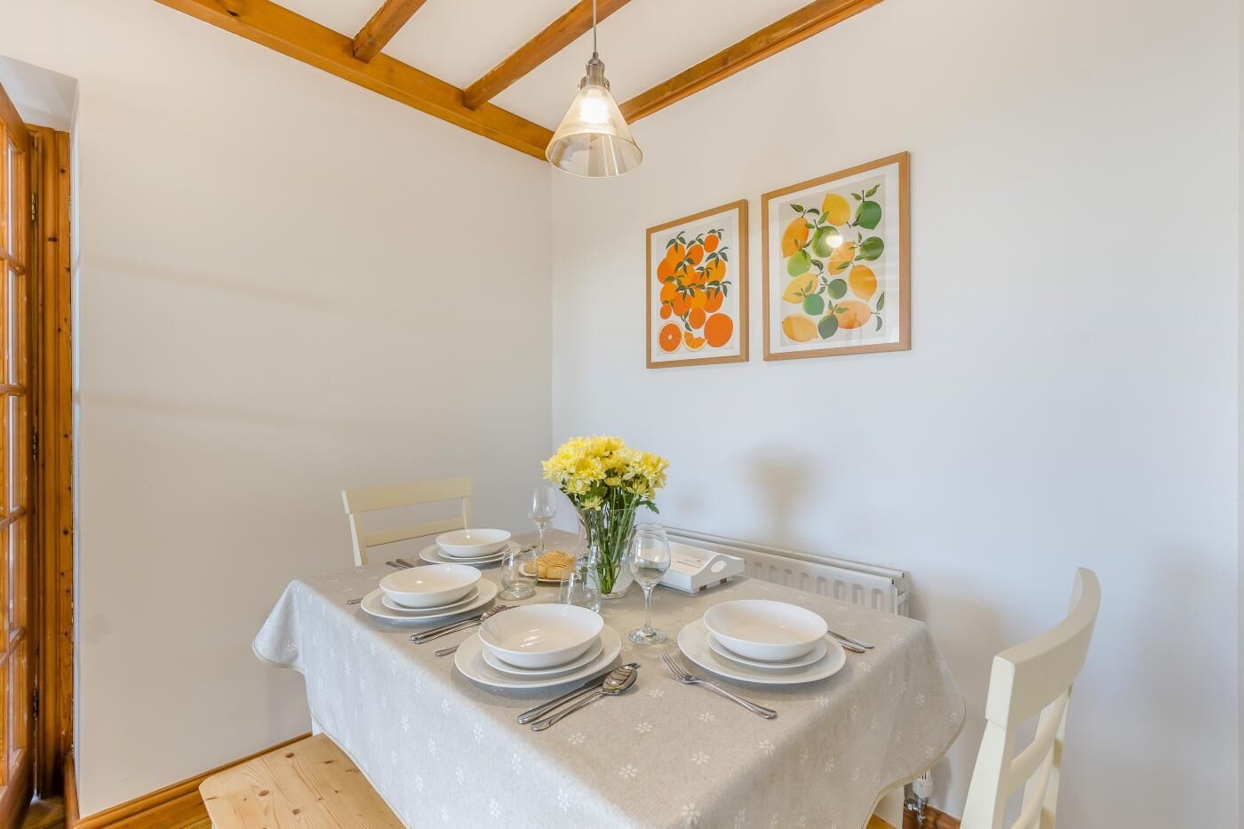 Un comedor con una mesa dispuesta para cuatro, sillas blancas, una lámpara colgante y dos obras de arte con motivos de frutas.