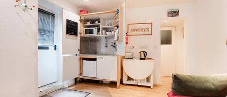 A compact kitchen with a microwave, sink, and storage cabinets. There is a small seating area with a green cushion and a red pillow.