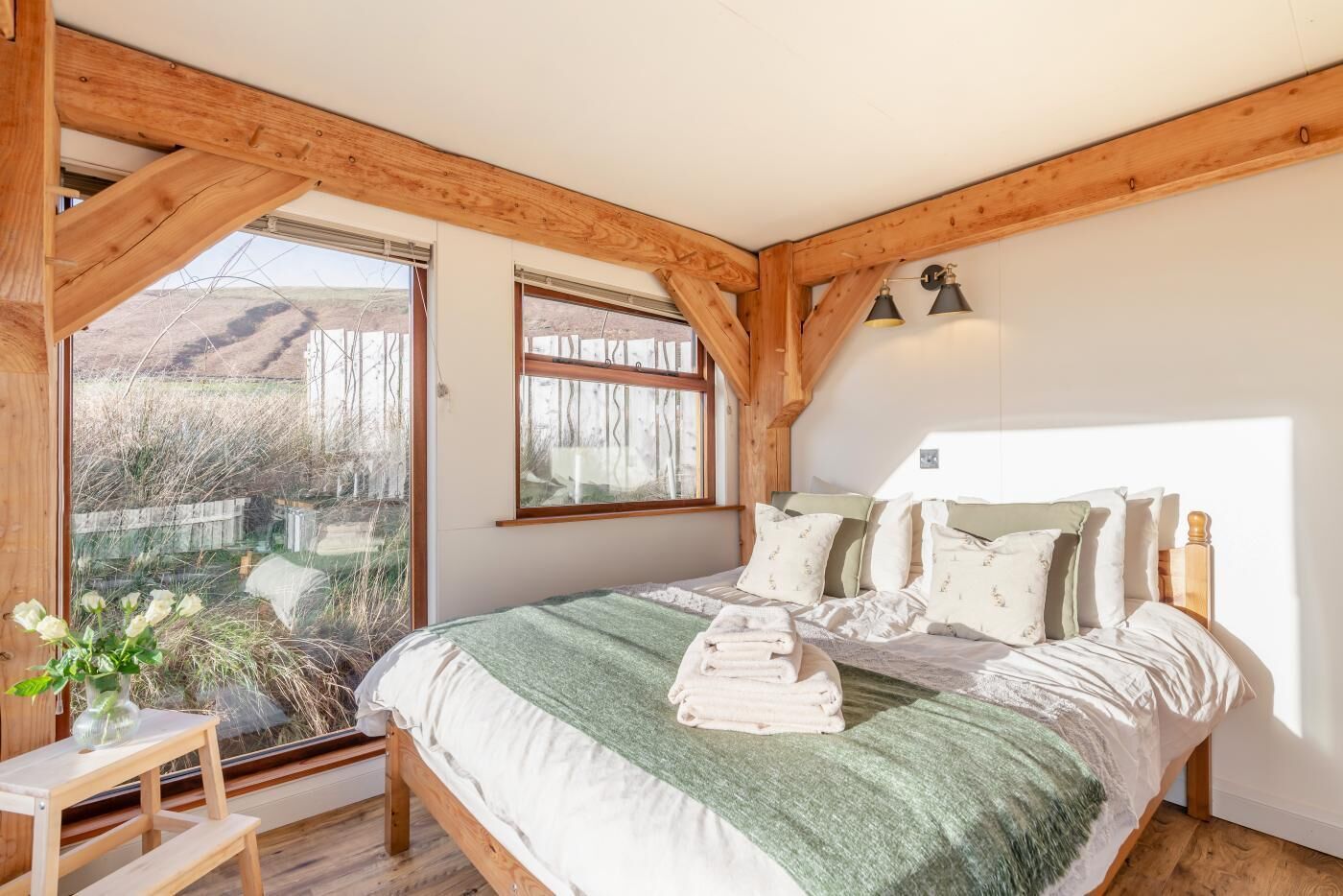 Une chambre à coucher avec un grand lit, des poutres en bois, une fenêtre donnant sur un beau paysage et une table de chevet agrémentée d’un vase.