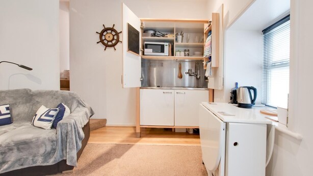 Un espacio habitable pequeño con kitchenette, un sofá con cojines, un mueble bajo y una ventana con persianas.