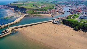 Vista aérea de un pueblo costero con playa de arena, puerto fortificado y campos verdes circundantes.