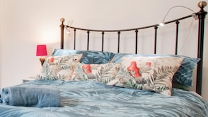 Una cama con colcha azul estampada con motivos florales, varias almohadas con diseños tropicales, un cabecero de madera y una lámpara de noche.