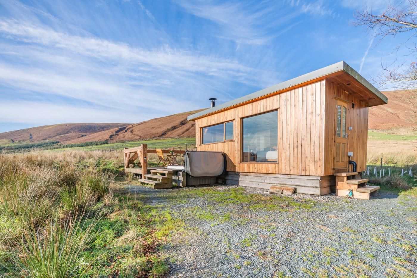 Une cabane en bois dotée de grandes fenêtres, d’une terrasse et d’un chemin de gravier.